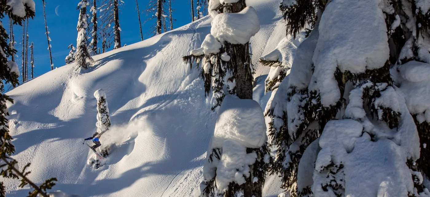 Skier mid-air during backcountry jump with snowy peaks backdrop